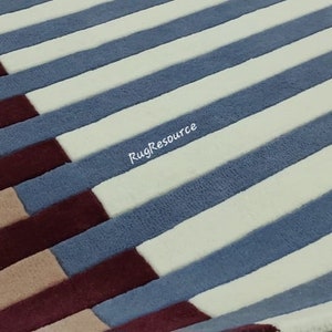 May include: A close-up of a striped rug with alternating bands of white, blue, burgundy, and beige. The rug has a soft, plush texture.