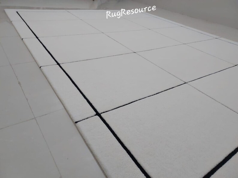Pu&ograve; includere: Tappeto rettangolare bianco con un design geometrico. Il tappeto presenta un motivo a griglia con linee scure. Il testo "RugResource" si trova nell'angolo in alto a destra. Il tappeto &egrave; su una superficie bianca.
