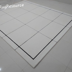 Pu&ograve; includere: Un tappeto bianco con un motivo a griglia, con linee nere che creano un design geometrico. Il tappeto ha un bordo nero e il testo "RugResource" nell'angolo in alto a sinistra.
