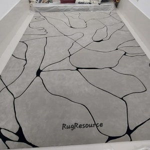 Puede incluir: Una alfombra de &aacute;rea abstracta gris y negra con un dise&ntilde;o moderno. La alfombra presenta un patr&oacute;n de l&iacute;neas negras que crean una serie de formas org&aacute;nicas sobre un fondo gris. La alfombra est&aacute; hecha de un material suave y mullido y es perfecta para agregar un toque de estilo moderno a cualquier habitaci&oacute;n.