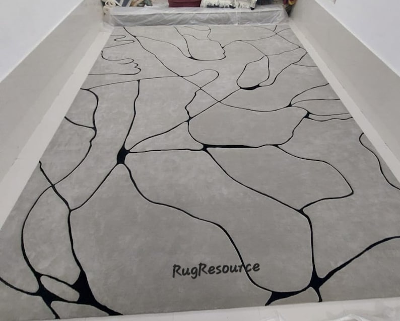 Puede incluir: Una alfombra de &aacute;rea abstracta gris y negra con un dise&ntilde;o moderno. La alfombra presenta un patr&oacute;n de l&iacute;neas negras que crean una serie de formas org&aacute;nicas sobre un fondo gris. La alfombra est&aacute; hecha de un material suave y mullido y es perfecta para agregar un toque de estilo moderno a cualquier habitaci&oacute;n.
