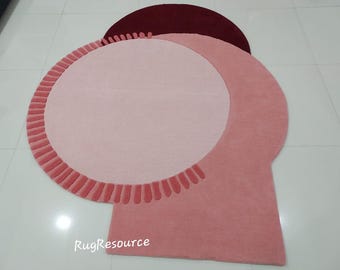 Puede incluir: Una alfombra redonda rosa y burdeos con un borde festoneado. La alfombra tiene un diseño único con un círculo más pequeño en el centro y un círculo más grande alrededor.