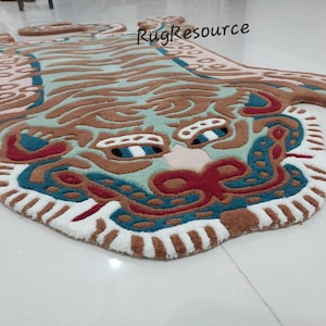 Op de afbeelding: Een decoratief tapijt in de vorm van een tijger. Het tapijt heeft een gedetailleerd ontwerp met bruine, turquoise, rode en witte kleuren. Het gezicht van de tijger is prominent aanwezig, met ingewikkelde patronen en een getextureerd uiterlijk. Het tapijt is ontworpen om op de vloer te worden geplaatst.