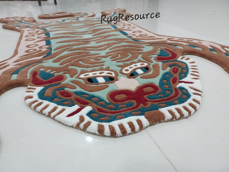 Op de afbeelding: Een decoratief tapijt in de vorm van een tijger. Het tapijt heeft een gedetailleerd ontwerp met bruine, turquoise, rode en witte kleuren. Het gezicht van de tijger is prominent aanwezig, met ingewikkelde patronen en een getextureerd uiterlijk. Het tapijt is ontworpen om op de vloer te worden geplaatst.