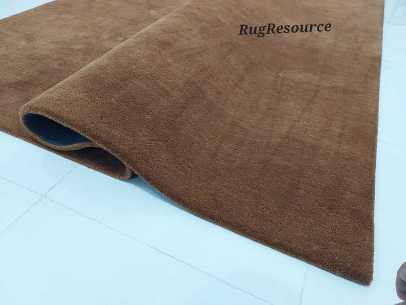 May include: Close-up of a brown, plush rug with a slightly darker brown border. The rug is rolled up and the edge is visible. The text "RugResource" is visible in the upper right corner of the image.