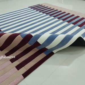 May include: A close-up of a blue, white, pink, and burgundy striped rug with a textured surface. The rug is folded in half, showing the underside of the rug.