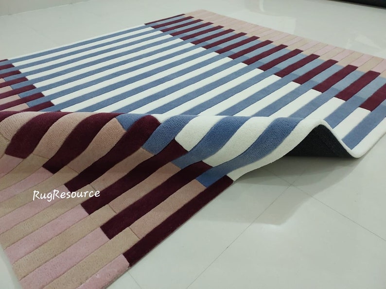 May include: A close-up of a blue, white, pink, and burgundy striped rug with a textured surface. The rug is folded in half, showing the underside of the rug.