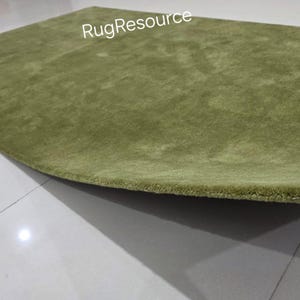 May include: A green rug with a soft, textured surface. The rug is a light shade of green and has a slightly curved shape. The edge of the rug is being held up, revealing its thickness. The text "RugResource" is visible.
