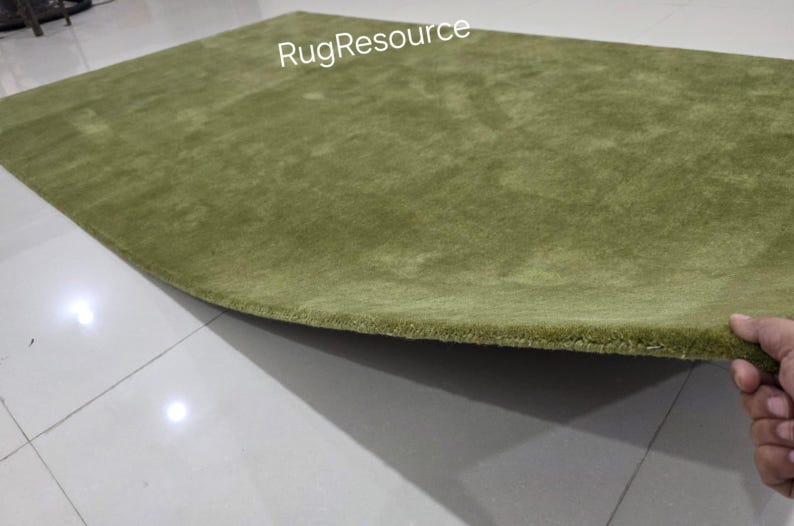 May include: A green rug with a soft, textured surface. The rug is a light shade of green and has a slightly curved shape. The edge of the rug is being held up, revealing its thickness. The text "RugResource" is visible.