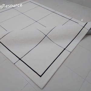 Pu&ograve; includere: Tappeto bianco con un design geometrico. Il tappeto presenta un motivo a griglia con linee nere e un bordo nero. L'angolo del tappeto &egrave; ripiegato, rivelando un retro grigio. Il testo "RugResource" si trova nell'angolo in alto a sinistra.