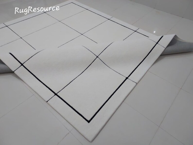 Pu&ograve; includere: Tappeto bianco con un design geometrico. Il tappeto presenta un motivo a griglia con linee nere e un bordo nero. L'angolo del tappeto &egrave; ripiegato, rivelando un retro grigio. Il testo "RugResource" si trova nell'angolo in alto a sinistra.