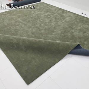 K&ouml;nnte beinhalten: Ein gro&szlig;er, olivgr&uuml;ner Teppich mit weicher, strukturierter Oberfl&auml;che. Der Teppich ist teilweise ausgerollt und zeigt eine dunkelblaue R&uuml;ckseite. Der Text "Rug Resource" ist sichtbar.