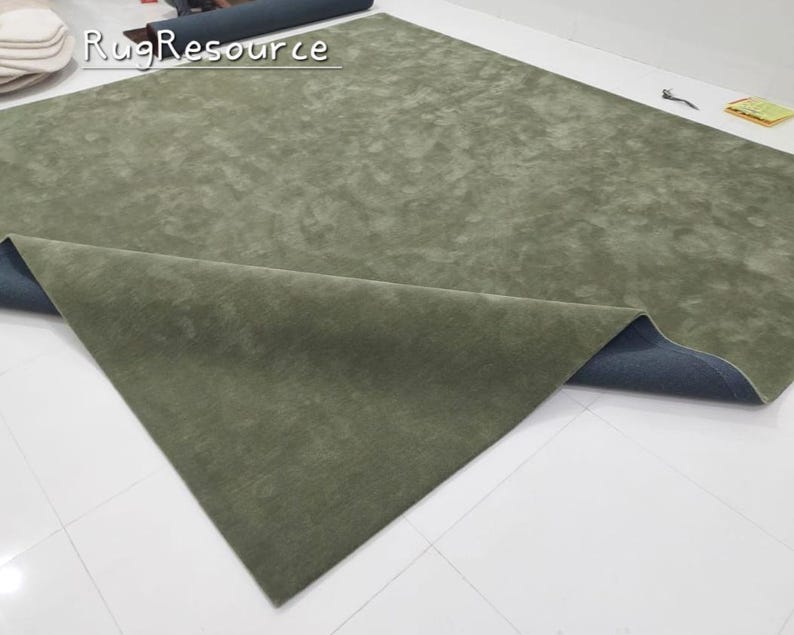 K&ouml;nnte beinhalten: Ein gro&szlig;er, olivgr&uuml;ner Teppich mit weicher, strukturierter Oberfl&auml;che. Der Teppich ist teilweise ausgerollt und zeigt eine dunkelblaue R&uuml;ckseite. Der Text "Rug Resource" ist sichtbar.