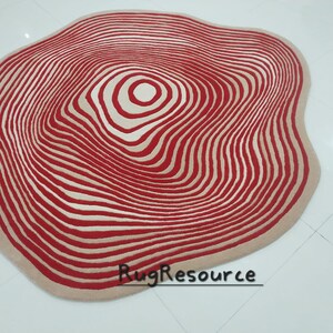 May include: A large, round, cream-colored rug with a red and cream concentric circle pattern. The rug has a wavy, organic shape.