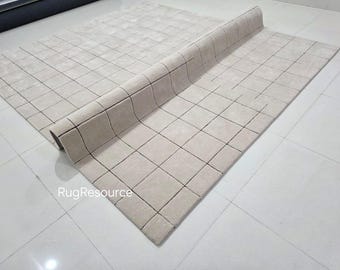 May include: Beige rectangular rug with a grid pattern. The rug is partially rolled up, revealing its thickness and construction. The grid lines are dark, contrasting with the light beige background. The rug is on a white tiled floor.