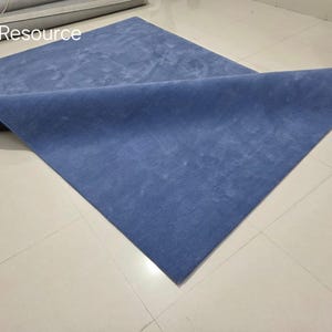 May include: A blue rectangular rug with a folded corner, displayed on a tiled floor. The rug has a soft, textured appearance and a solid color. The text "RugResource" is visible in the upper left corner.
