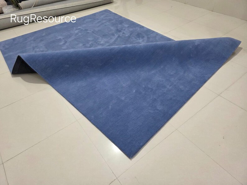 May include: A blue rectangular rug with a folded corner, displayed on a tiled floor. The rug has a soft, textured appearance and a solid color. The text "RugResource" is visible in the upper left corner.