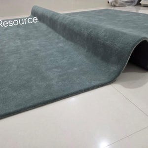 K&ouml;nnte beinhalten: Ein rechteckiger, teal-farbener Teppich mit weicher, strukturierter Oberfl&auml;che, teilweise aufgerollt. Die dunkle Unterseite des Teppichs ist sichtbar. Die Worte "RugResource" sind in der oberen linken Ecke. Der Teppich liegt auf einem wei&szlig;en Fliesenboden.