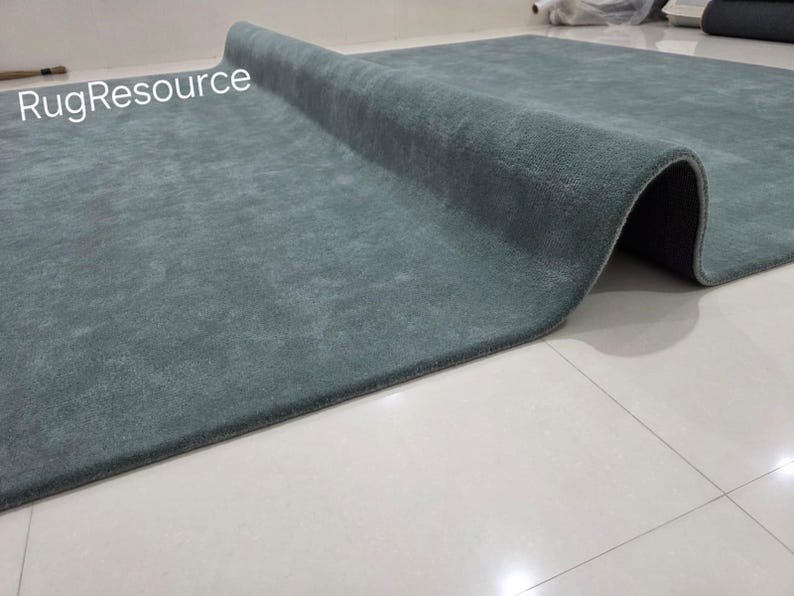 K&ouml;nnte beinhalten: Ein rechteckiger, teal-farbener Teppich mit weicher, strukturierter Oberfl&auml;che, teilweise aufgerollt. Die dunkle Unterseite des Teppichs ist sichtbar. Die Worte "RugResource" sind in der oberen linken Ecke. Der Teppich liegt auf einem wei&szlig;en Fliesenboden.