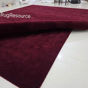 K&ouml;nnte beinhalten: Ein gro&szlig;er, rechteckiger burgunderfarbener Teppich mit einer weichen, strukturierten Oberfl&auml;che. Der Teppich ist teilweise aufgerollt und gibt die Unterseite frei. Der Text "RugResource" ist in der oberen linken Ecke sichtbar. Der Teppich liegt auf einem wei&szlig;en Fliesenboden.