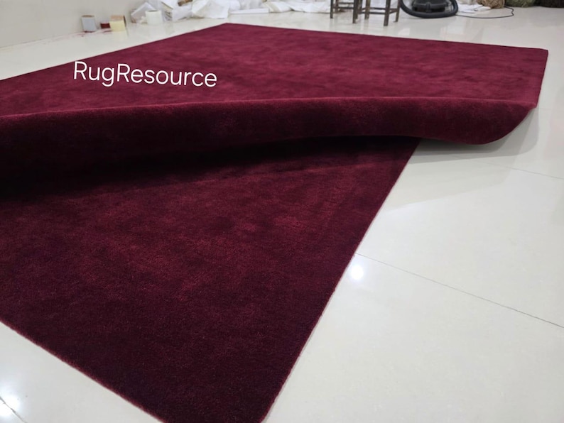 K&ouml;nnte beinhalten: Ein gro&szlig;er, rechteckiger burgunderfarbener Teppich mit einer weichen, strukturierten Oberfl&auml;che. Der Teppich ist teilweise aufgerollt und gibt die Unterseite frei. Der Text "RugResource" ist in der oberen linken Ecke sichtbar. Der Teppich liegt auf einem wei&szlig;en Fliesenboden.