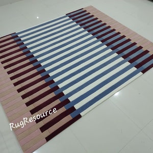 May include: A striped rug with a geometric pattern in shades of pink, burgundy, and blue. The rug has a modern design and is made of soft, plush material. The text "RugResource" is visible in the lower left corner of the rug.