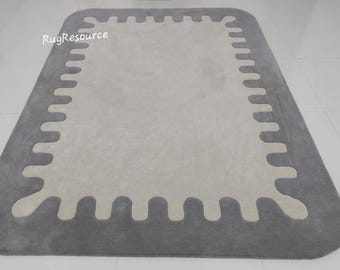May include: A grey and white area rug with a unique, irregular border design. The rug has a light grey border with a white, scalloped edge.