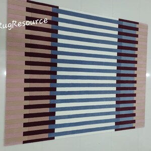 May include: A striped rug with a geometric pattern in shades of pink, burgundy, blue, and white. The rug has a modern design and is perfect for adding a touch of color and style to any room.