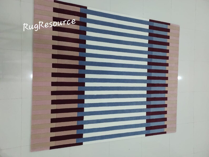 May include: A striped rug with a geometric pattern in shades of pink, burgundy, blue, and white. The rug has a modern design and is perfect for adding a touch of color and style to any room.