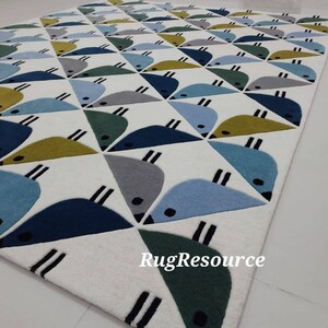 May include: A white rug with a geometric pattern of blue, green, and yellow birds. The rug has a white background with a black outline around the birds. The rug is made of a soft, plush material. The text "RugResource" is printed in the bottom right corner of the rug.