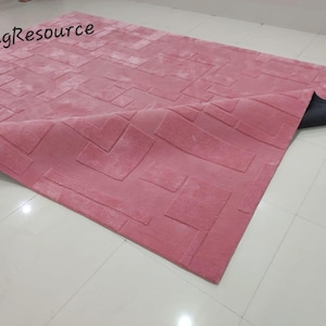 May include: A pink rug with a geometric pattern. The rug is made of a soft, plush material and has a subtle sheen. The rug is folded in half, revealing the black backing.