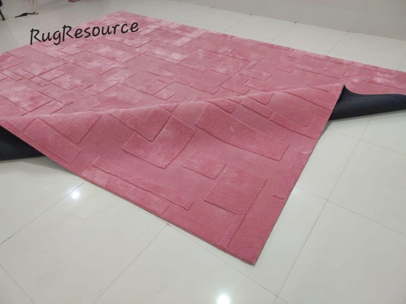 May include: A pink rug with a geometric pattern. The rug is made of a soft, plush material and has a subtle sheen. The rug is folded in half, revealing the black backing.