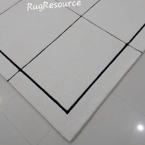 Pu&ograve; includere: Primo piano di un tappeto bianco con un design geometrico. Il tappeto presenta un motivo a griglia con bordi neri. Il testo "RugResource" &egrave; visibile nell'angolo in alto a sinistra. Il tappeto &egrave; su un pavimento piastrellato.