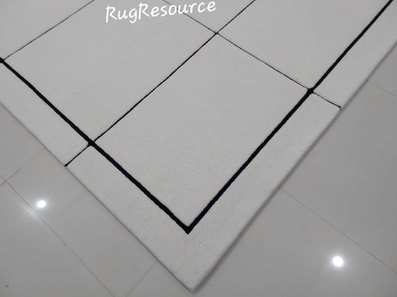 Pu&ograve; includere: Primo piano di un tappeto bianco con un design geometrico. Il tappeto presenta un motivo a griglia con bordi neri. Il testo "RugResource" &egrave; visibile nell'angolo in alto a sinistra. Il tappeto &egrave; su un pavimento piastrellato.