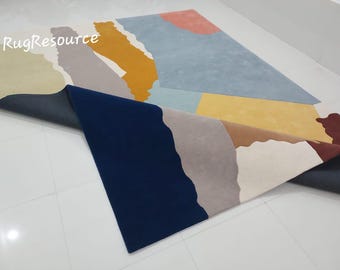 Puede incluir: Una alfombra abstracta colorida con formas geométricas en tonos de azul, amarillo, marrón, rosa y blanco. La alfombra está doblada por la mitad, mostrando el reverso, que es de color azul oscuro.
