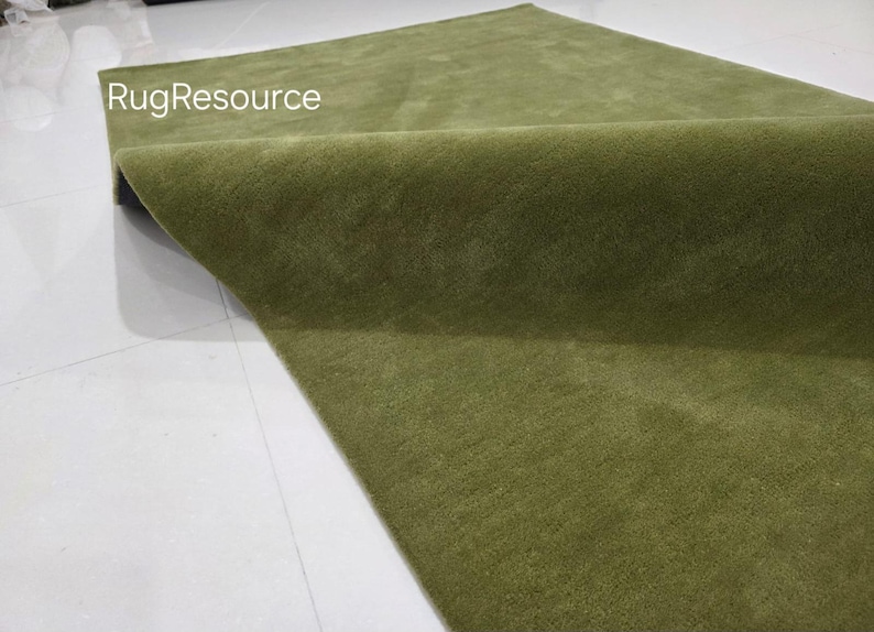 May include: A folded, olive green rug with a soft, textured surface. The rug is displayed on a white surface, with the text "RugResource" visible in the upper left corner. The rug's color is consistent throughout.