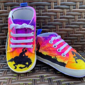 Puede incluir: Un par de zapatos de bebé con un diseño de puesta de sol y silueta de vaquero. Los zapatos están pintados con un degradado de rosa, naranja y amarillo. El vaquero está montando un caballo.