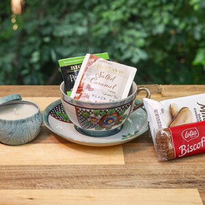 May include: A teacup and saucer with a floral design, filled with tea bags, a teal blue candle in a tin, and a package of Lotus Biscoff cookies.