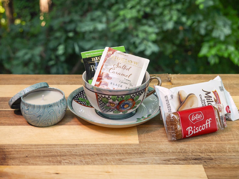 May include: A teacup and saucer with a floral design, filled with tea bags, a teal blue candle in a tin, and a package of Lotus Biscoff cookies.