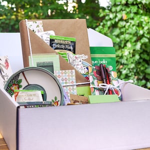 May include: A white box filled with a variety of items, including a brown notebook, a green and white tea bag, a green and white patterned plate, a green and white patterned notepad, a green and white patterned sticker sheet, a green and white patterned pen, and a green and white patterned envelope. The box is sitting on a wooden surface.