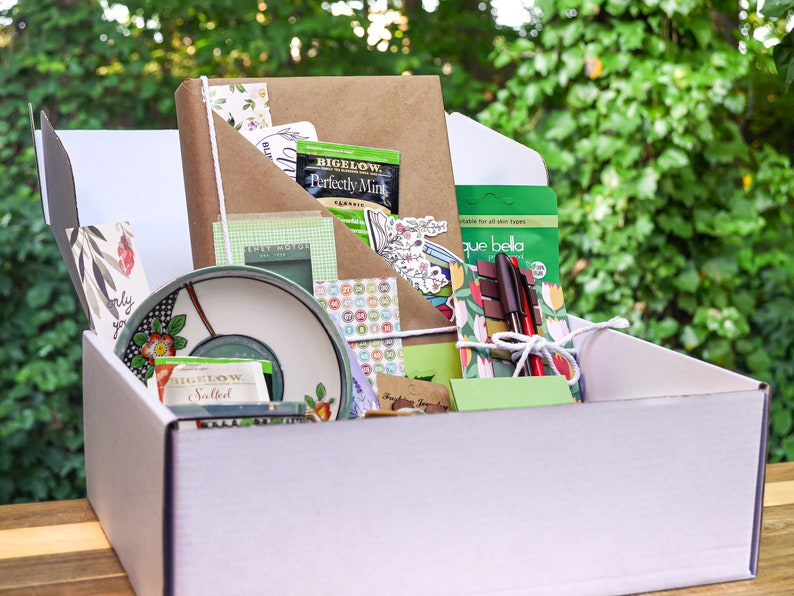May include: A white box filled with a variety of items, including a brown notebook, a green and white tea bag, a green and white patterned plate, a green and white patterned notepad, a green and white patterned sticker sheet, a green and white patterned pen, and a green and white patterned envelope. The box is sitting on a wooden surface.