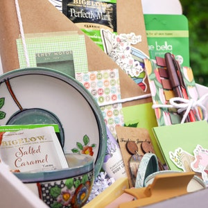 May include: A white gift box filled with various items, including tea bags, a teacup and saucer, stationery, stickers, and jewelry. The box is decorated with a green and white floral pattern.