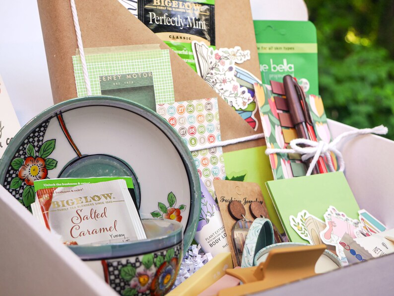May include: A white gift box filled with various items, including tea bags, a teacup and saucer, stationery, stickers, and jewelry. The box is decorated with a green and white floral pattern.