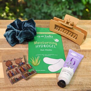 May include: A flat lay of various beauty and fashion items on a wooden surface. The items include a teal velvet scrunchie, a brown hair clip, a package of hydrogel eye masks, a tube of lavender body lotion, and a pair of book-shaped earrings.