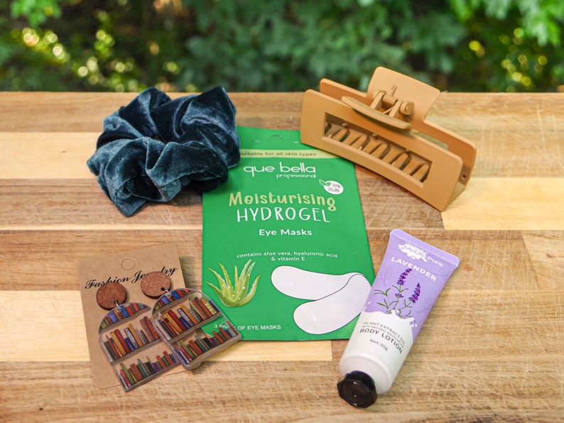 May include: A flat lay of various beauty and fashion items on a wooden surface. The items include a teal velvet scrunchie, a brown hair clip, a package of hydrogel eye masks, a tube of lavender body lotion, and a pair of book-shaped earrings.
