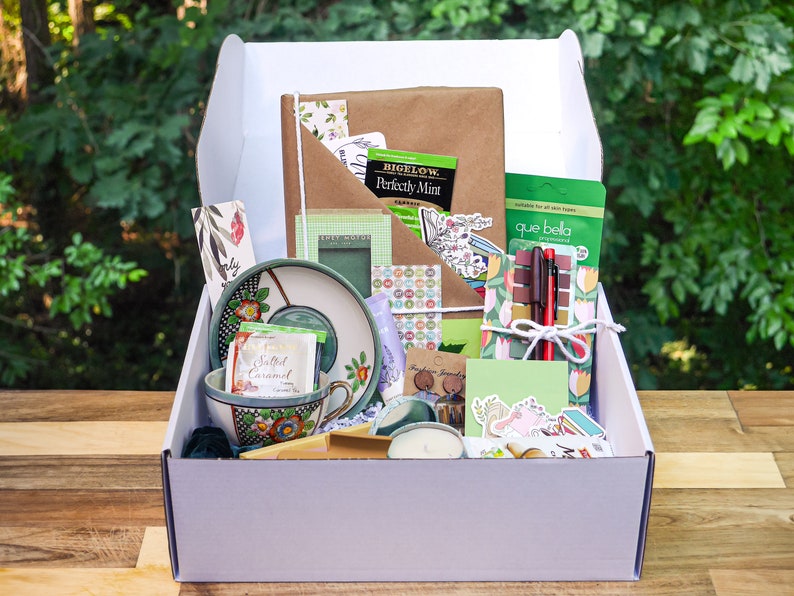May include: A white box filled with a variety of items, including a teacup and saucer, a notebook, a candle, pens, and stickers. The box is open and the items are visible. The box is sitting on a wooden surface.