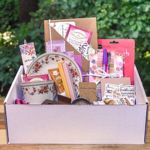 May include: A white box filled with a variety of items, including a teacup, tea bags, a small plate, a notebook, pens, stickers, and a face mask. The box is on a wooden surface.