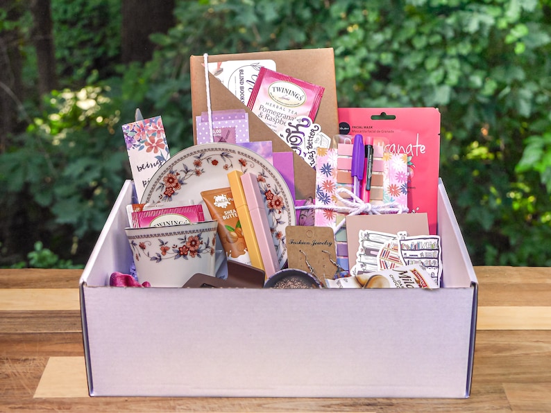 May include: A white box filled with a variety of items, including a teacup, tea bags, a small plate, a notebook, pens, stickers, and a face mask. The box is on a wooden surface.