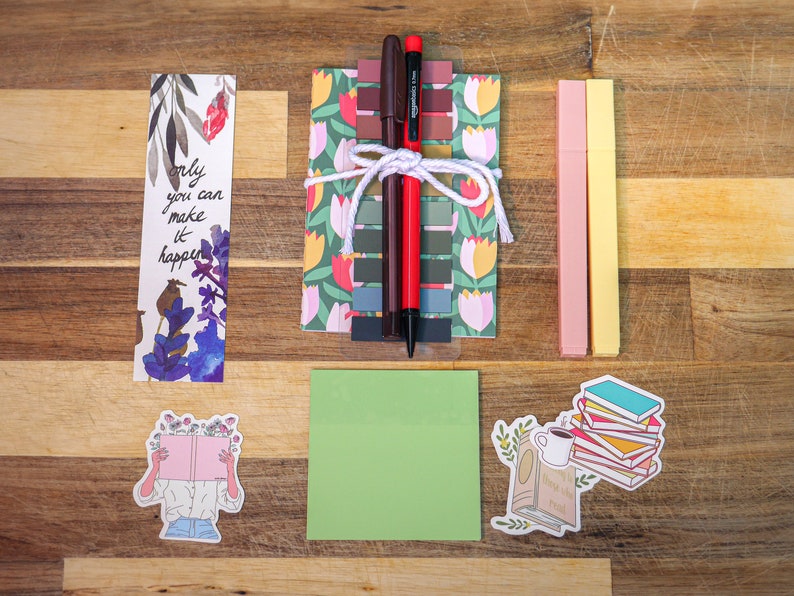 May include: A set of stationery supplies including two bookmarks, two highlighters, a set of sticky notes, a green notepad, and two stickers. One bookmark features a floral design and the text "only you can make it happen". One sticker features a person reading a book with flowers on top of the book. The other sticker features a stack of books with a cup of coffee and a plant.
