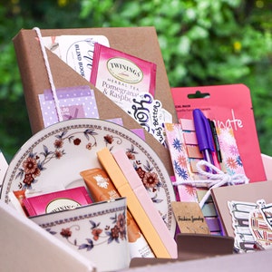 May include: A pink and white gift box filled with various items, including a brown notebook, a tea bag, a pink and purple sticker, a floral patterned pouch, a purple pen, a pink and white patterned notepad, a pink and white patterned bookmark, a teacup and saucer, and a pink and white patterned sticker. The box is surrounded by green foliage.